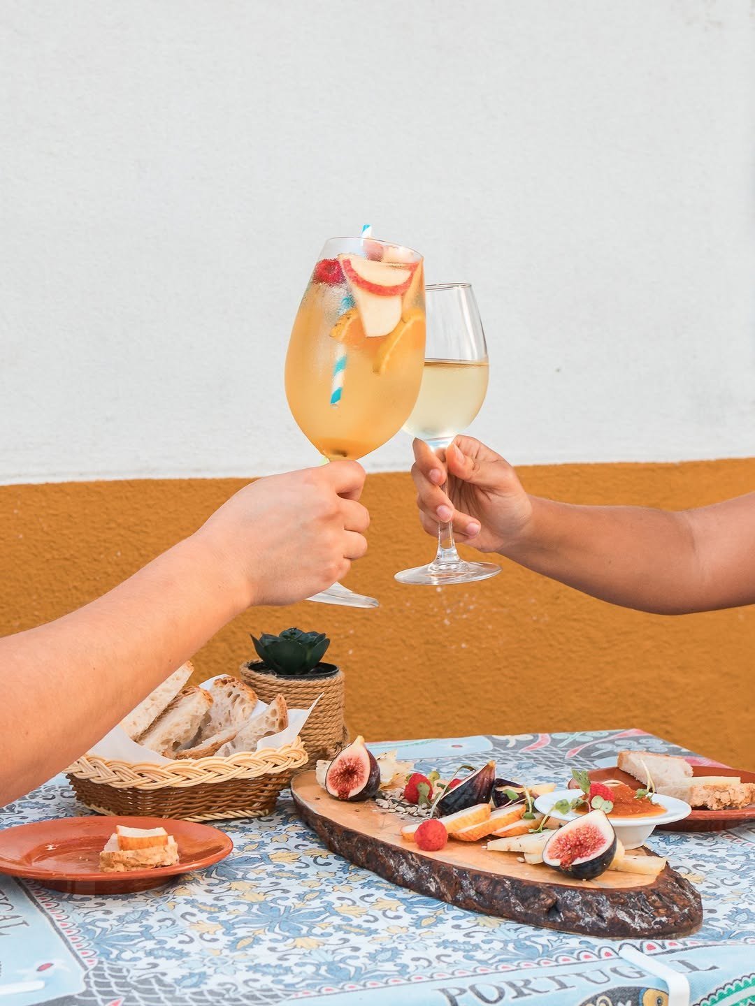 Dois copos a brindar sobre uma mesa com tábua de queijos e figos, junto a uma parede branca e amarela